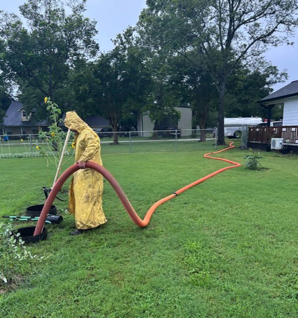 A OnePump worker in a full-length hooded protective suit holding the suction hose over an open septic tank, hose running across the lawn to the truck