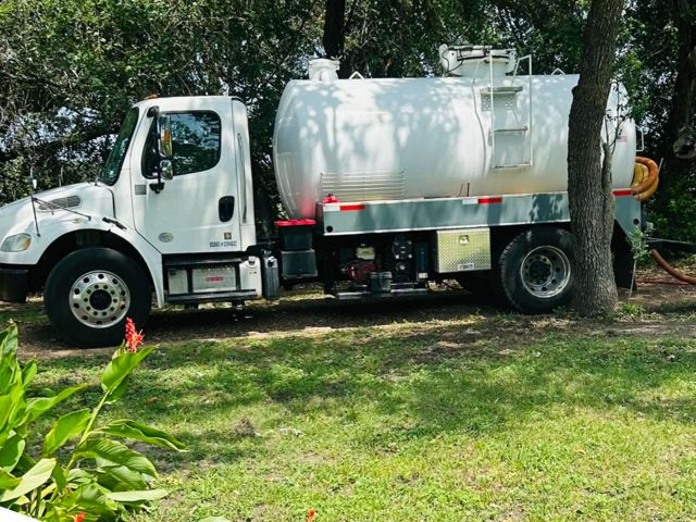 The white OnePump vacuum truck parked in the shade of trees on a rural property, suction hose running out to the job